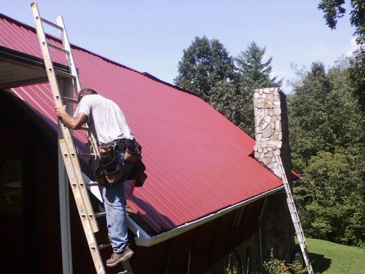 Roofer on a ladder beside a red metal roof and stone chimney — Soffit & Fascia Repair by Roofing Brothers in Saint Peter